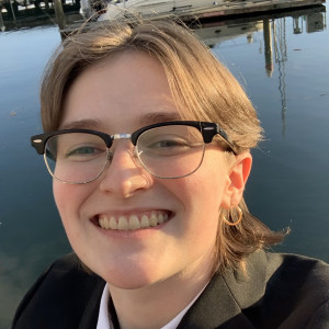 Devyn, wearing glasses, a white collared shirt with a black jacket and a gold broach, smiles in front of a marina scene with water in the background.