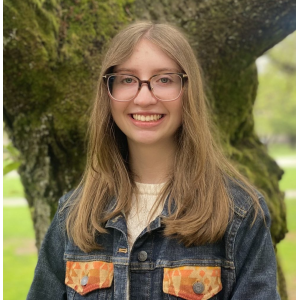 Olivia, long hair and glasses in a jean jacket, stands in front of a cherry tree.