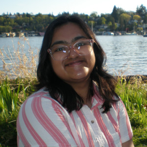 Sruti, wearing glasses and a pink and white striped shirt, smiles on a grassy lawn with water behind her.