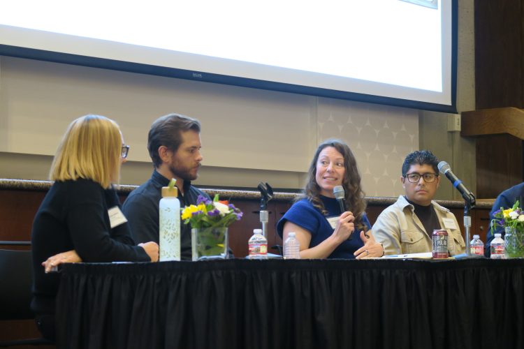 A panel of people sit around a table, one person hold a microphone speaking.