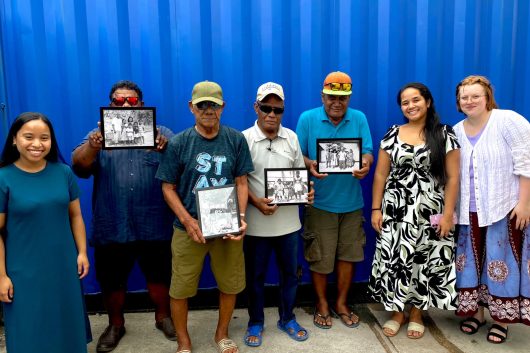 Seven people stand in front of a blue wall, some hold framed photos.