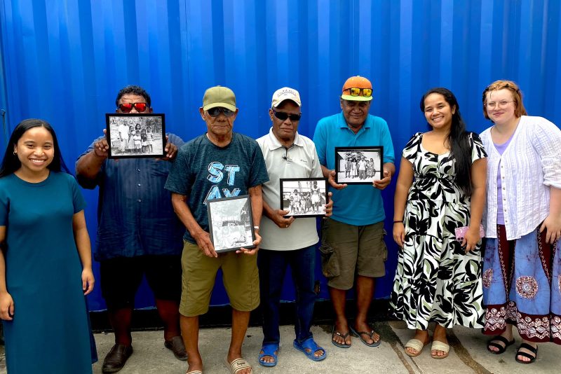 Seven people stand in front of a blue wall, some hold framed photos.