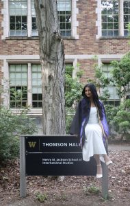 Sejal Antal in graduation regalia sitting on the building sign in front of Thomson Hall at the University of Washington.