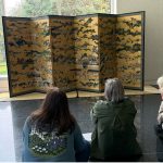 Three people sit in front of a six-panel screen at the Seattle Asian Art Museum.