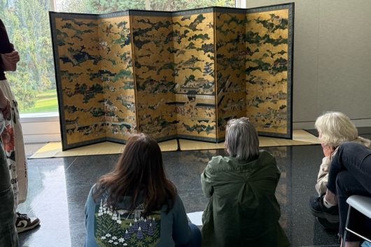 Three people sit in front of a six-panel screen at the Seattle Asian Art Museum.