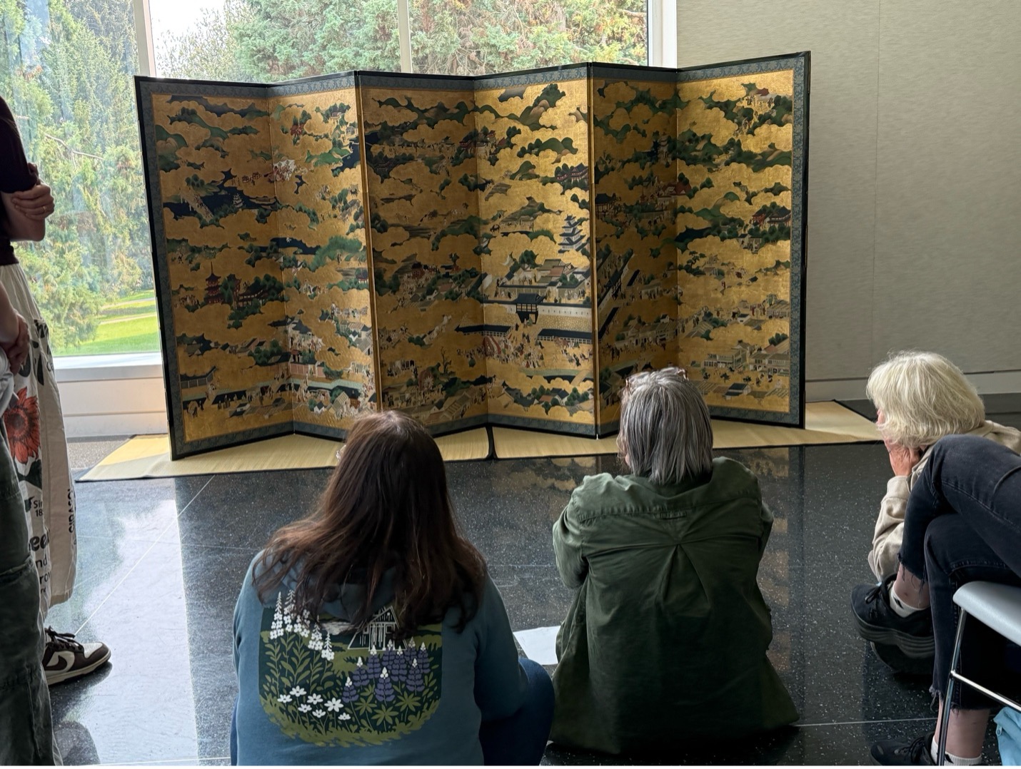 Three people sit in front of a six-panel screen at the Seattle Asian Art Museum.